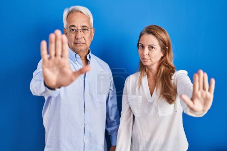 Photo for Middle age hispanic couple standing over blue background doing stop sing with palm of the hand. warning expression with negative and serious gesture on the face. - Royalty Free Image