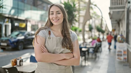 Foto de Joven hermosa mujer hispana camarera sonriendo confiada de pie con los brazos cruzados gesto en la terraza de la cafetería - Imagen libre de derechos