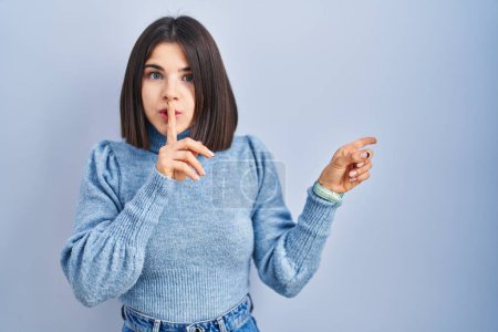 Téléchargez les photos : Young hispanic woman standing over blue background asking to be quiet with finger on lips pointing with hand to the side. silence and secret concept. - en image libre de droit