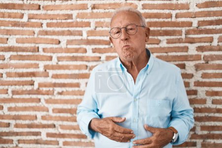 Foto de Senior man with grey hair standing over bricks wall puffing cheeks with funny face. mouth inflated with air, crazy expression. - Imagen libre de derechos