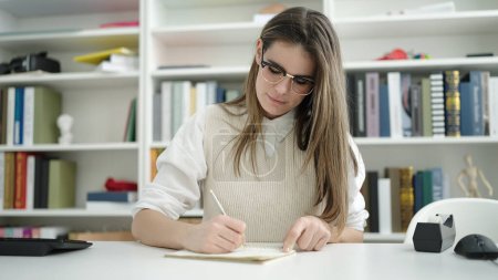 Téléchargez les photos : Jeune belle étudiante hispanique écrivant sur carnet à la bibliothèque - en image libre de droit