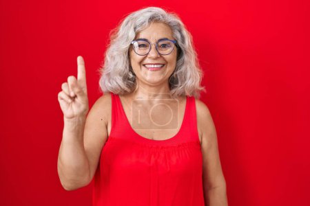 Téléchargez les photos : Femme d'âge moyen avec les cheveux gris debout sur fond rouge montrant et pointant vers le haut avec le doigt numéro un tout en souriant confiant et heureux. - en image libre de droit