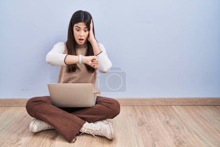 Téléchargez les photos : Jeune femme brune travaillant à l'aide d'un ordinateur portable assis sur le sol en regardant le temps de la montre inquiet, peur de se faire tard - en image libre de droit