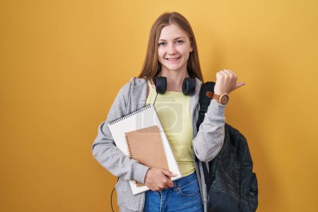 Téléchargez les photos : Jeune femme caucasienne portant un sac à dos étudiant et tenant des livres pointant vers le dos avec la main et les pouces levés, souriant confiant - en image libre de droit