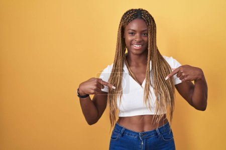 Téléchargez les photos : Femme afro-américaine avec les cheveux tressés debout sur fond jaune regardant confiant avec le sourire sur le visage, se montrant du doigt fier et heureux. - en image libre de droit