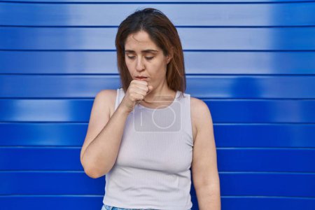 Foto de Mujer morena parada sobre fondo azul sintiéndose mal y tosiendo como síntoma de resfriado o bronquitis. concepto de atención sanitaria. - Imagen libre de derechos