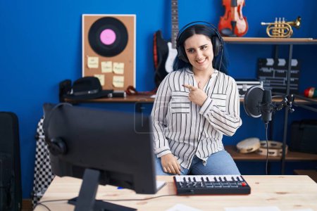 Téléchargez les photos : Jeune fille moderne avec les cheveux bleus au studio de musique portant des écouteurs souriant joyeuse pointant avec la main et le doigt sur le côté - en image libre de droit