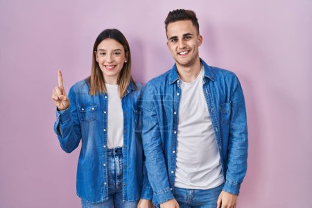 Téléchargez les photos : Jeune couple hispanique debout sur fond rose montrant et pointant vers le haut avec le doigt numéro un tout en souriant confiant et heureux. - en image libre de droit