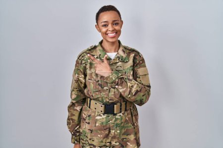 Foto de Hermosa mujer afroamericana vistiendo camuflaje uniforme del ejército alegre con una sonrisa en la cara señalando con la mano y el dedo hacia el lado con expresión feliz y natural - Imagen libre de derechos