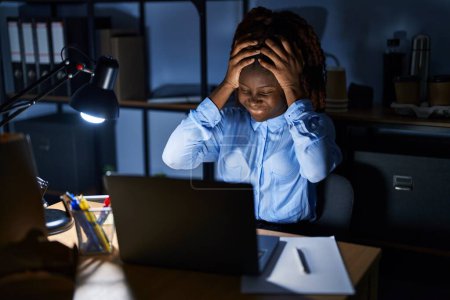 Photo for African woman working at the office at night suffering from headache desperate and stressed because pain and migraine. hands on head. - Royalty Free Image