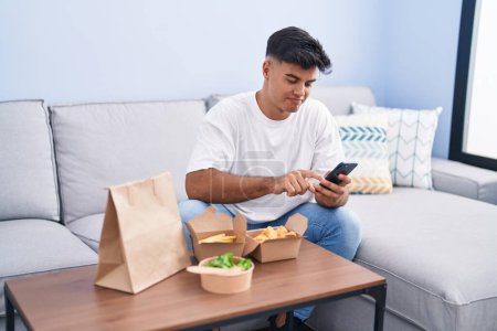 Téléchargez les photos : Jeune homme hispanique manger emporter de la nourriture à l'aide d'un smartphone à la maison - en image libre de droit