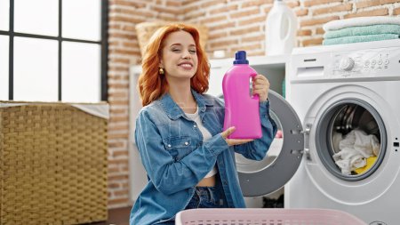 Photo for Young redhead woman washing clothes holding detergent bottle at laundry room - Royalty Free Image