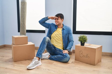 Photo for Young hispanic man sitting on the floor at new home very happy and smiling looking far away with hand over head. searching concept. - Royalty Free Image