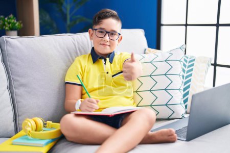 Photo for Young hispanic kid doing homework sitting on the sofa approving doing positive gesture with hand, thumbs up smiling and happy for success. winner gesture. - Royalty Free Image