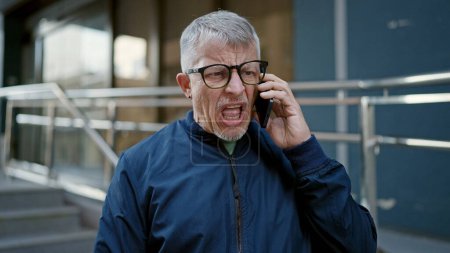 Foto de Hombre de pelo gris de mediana edad de pie con expresión furiosa hablando en el teléfono inteligente en la calle - Imagen libre de derechos