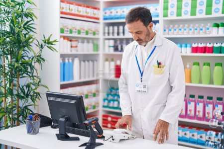 Téléchargez les photos : Pharmacien d'âge moyen tenant le téléphone en utilisant l'ordinateur à la pharmacie - en image libre de droit