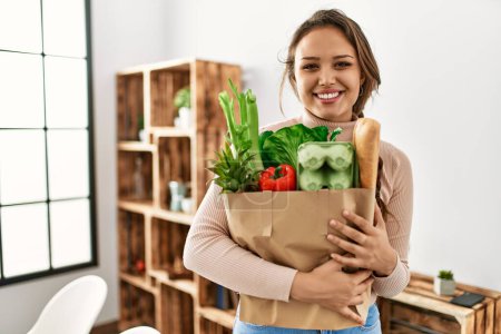 Téléchargez les photos : Jeune belle femme hispanique souriant confiant tenant sac d'épicerie à la maison - en image libre de droit