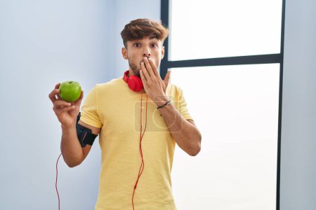 Photo for Arab man with beard wearing sportswear eating green apple covering mouth with hand, shocked and afraid for mistake. surprised expression - Royalty Free Image