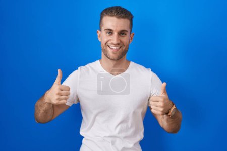 Téléchargez les photos : Jeune homme caucasien debout sur fond bleu signe de succès faisant geste positif avec la main, pouces levés souriant et heureux. expression joyeuse et geste gagnant. - en image libre de droit