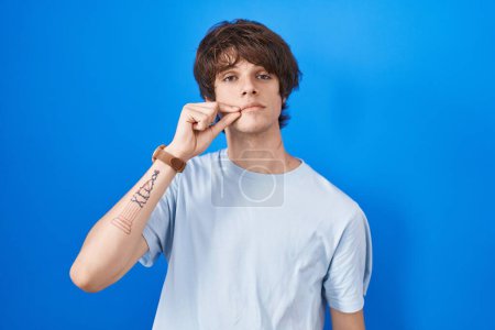 Téléchargez les photos : Jeune homme hispanique debout sur fond bleu bouche et lèvres fermées comme zip avec les doigts. secret et silencieux, tabou parlant - en image libre de droit