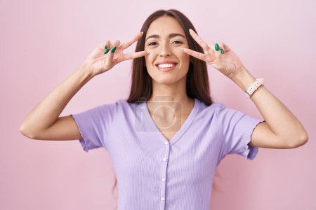 Foto de Mujer hispana joven con el pelo largo de pie sobre fondo rosa haciendo símbolo de paz con los dedos sobre la cara, sonriendo alegre mostrando la victoria - Imagen libre de derechos