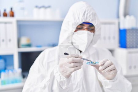 Foto de Joven científico hispano con máscara médica sosteniendo muestra con pinzas en laboratorio - Imagen libre de derechos
