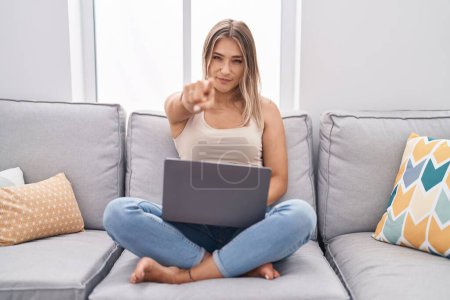 Photo for Blonde caucasian woman using laptop at home sitting on the sofa pointing with finger to the camera and to you, confident gesture looking serious - Royalty Free Image