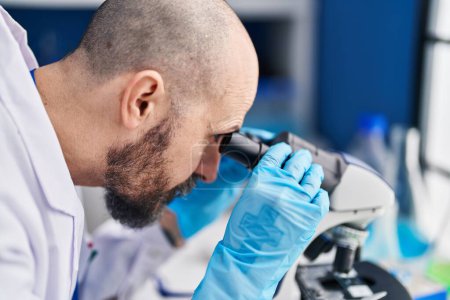 Téléchargez les photos : Jeune homme chauve scientifique utilisant un microscope en laboratoire - en image libre de droit