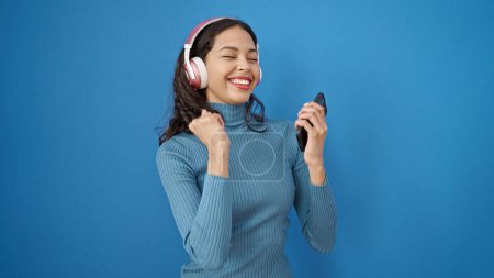 Foto de Joven hermosa mujer hispana bailando usando auriculares usando teléfono inteligente sobre fondo azul aislado - Imagen libre de derechos