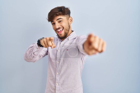 Téléchargez les photos : Homme arabe avec la barbe debout sur fond bleu pointant vers vous et la caméra avec les doigts, souriant positif et joyeux - en image libre de droit