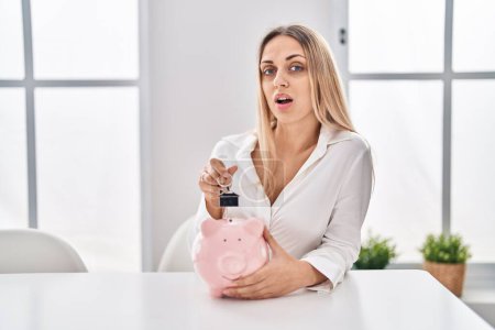 Photo for Young blonde woman holding piggy bank and house keys clueless and confused expression. doubt concept. - Royalty Free Image
