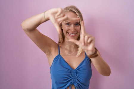 Foto de Mujer caucásica joven de pie sobre fondo rosa sonriendo haciendo marco con las manos y los dedos con la cara feliz. concepto de creatividad y fotografía. - Imagen libre de derechos