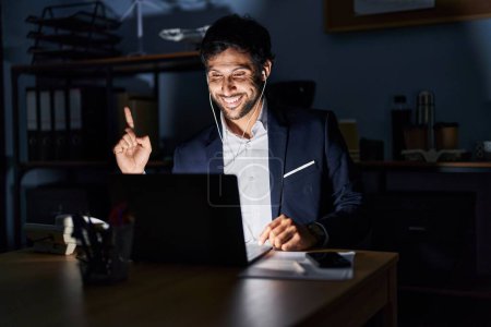 Téléchargez les photos : Un bel homme latin travaillant au bureau la nuit pointant du doigt avec une idée réussie. Sorti et heureux. numéro un. - en image libre de droit