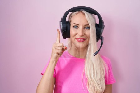 Téléchargez les photos : Femme caucasienne écouter de la musique à l'aide d'écouteurs montrant et pointant vers le haut avec le doigt numéro un tout en souriant confiant et heureux. - en image libre de droit