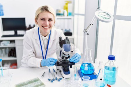 Téléchargez les photos : Jeune femme blonde scientifique utilisant un microscope souriant au laboratoire - en image libre de droit