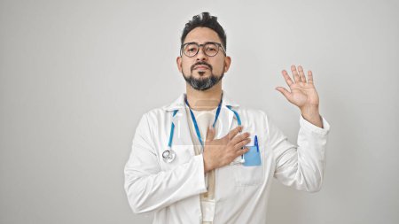 Foto de Joven médico latino haciendo un juramento con la mano en el pecho sobre un fondo blanco aislado - Imagen libre de derechos