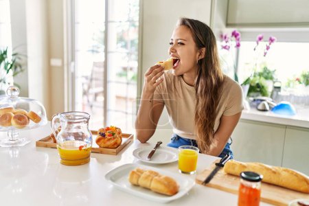 Photo for Young beautiful hispanic woman eating bread with jam having breakfast at the kitchen - Royalty Free Image