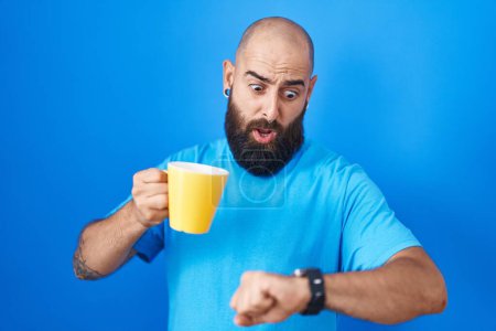 Téléchargez les photos : Jeune homme hispanique avec barbe et tatouages boire une tasse de café en regardant l'heure de la montre inquiet, peur de se retrouver en retard - en image libre de droit