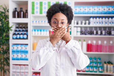 Téléchargez les photos : Belle femme africaine avec les cheveux bouclés travaillant à la pharmacie pharmacie choqué couvrant la bouche avec les mains pour erreur. concept secret. - en image libre de droit