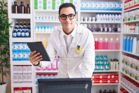 Photo for Young hispanic man pharmacist using touchpad and computer at pharmacy - Royalty Free Image