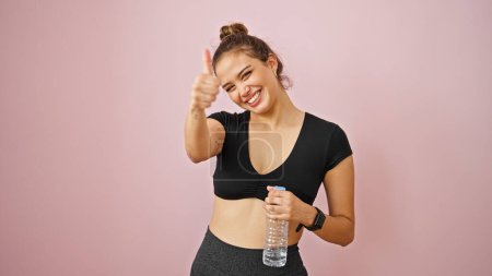 Foto de Joven mujer hispana hermosa usando ropa deportiva sosteniendo botella de agua haciendo gesto de pulgar hacia arriba sobre fondo rosa aislado - Imagen libre de derechos