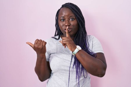 Téléchargez les photos : Jeune femme africaine debout sur fond rose demandant à être calme avec le doigt sur les lèvres pointant avec la main sur le côté. silence et concept secret. - en image libre de droit