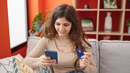Foto de Joven hermosa mujer hispana de compras con teléfono inteligente y tarjeta de crédito sentado en el sofá en casa - Imagen libre de derechos