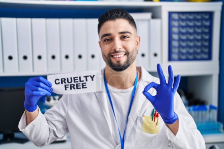 Foto de Young handsome man working at scientist laboratory holding cruelty free banner doing ok sign with fingers, smiling friendly gesturing excellent symbol - Imagen libre de derechos