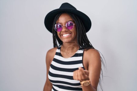 Foto de Joven afroamericano con trenzas con sombrero y gafas de sol haciendo señas ven aquí gesto con la mano invitando a la bienvenida feliz y sonriente - Imagen libre de derechos