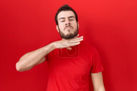 Téléchargez les photos : Jeune homme hispanique portant un t-shirt rouge décontracté tranchant la gorge avec la main comme couteau, menace d'agression avec une violence furieuse - en image libre de droit