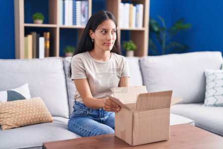 Photo for Young hispanic woman opening cardboard box smiling looking to the side and staring away thinking. - Royalty Free Image