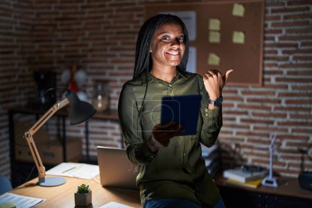 Téléchargez les photos : Jeune afro-américain avec des tresses travaillant au bureau la nuit pointant le dos derrière avec la main et les pouces levés, souriant confiant - en image libre de droit
