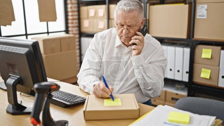 Téléchargez les photos : Middle age grey-haired man ecommerce business worker talking on smartphone writing on package at office - en image libre de droit