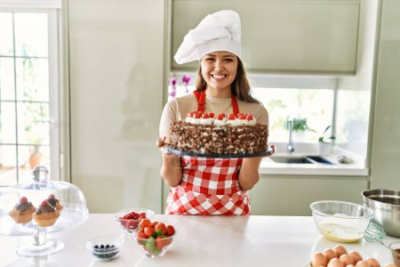 Téléchargez les photos : Jeune belle femme hispanique souriant confiant tenant gâteau à la cuisine - en image libre de droit